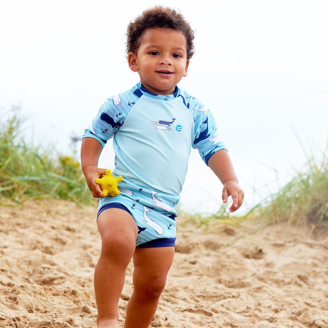 Lifestyle image of baby wearing a Happy Nappy Sunsuit in light blue and whales themed print. Front.