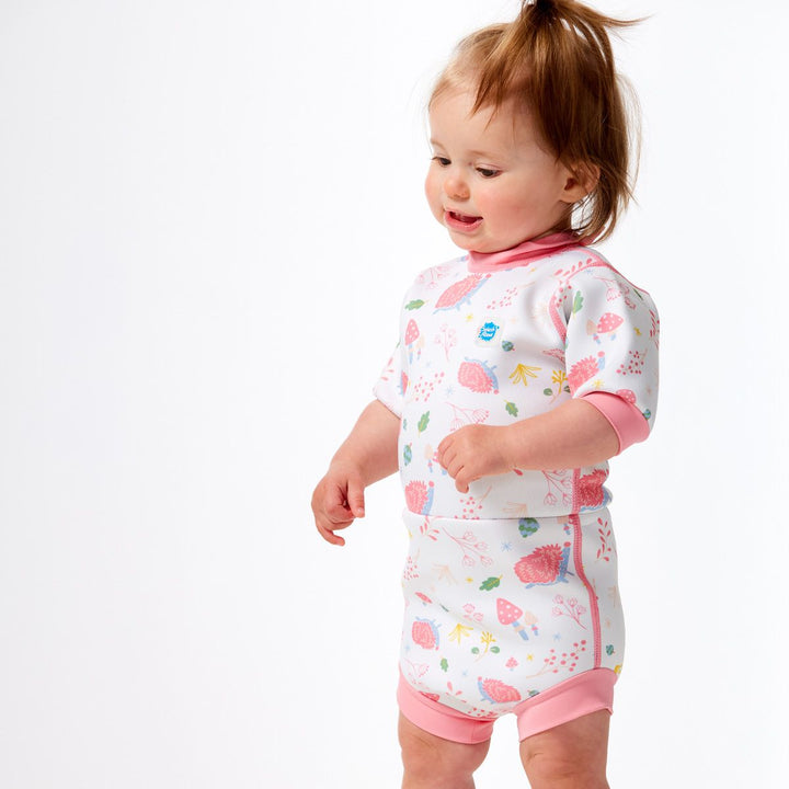 Lifestyle image of child wearing a baby wetsuit with built in swim nappy in white with pink trims and forest themed print, including hedgehogs, mushrooms and leaves. Side.