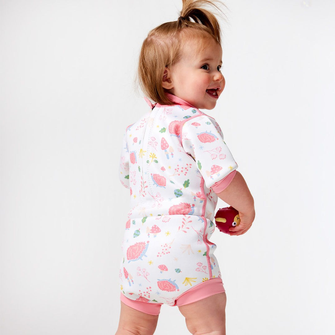 Lifestyle image of child wearing a baby wetsuit with built in swim nappy in white with pink trims and forest themed print, including hedgehogs, mushrooms and leaves. Back.