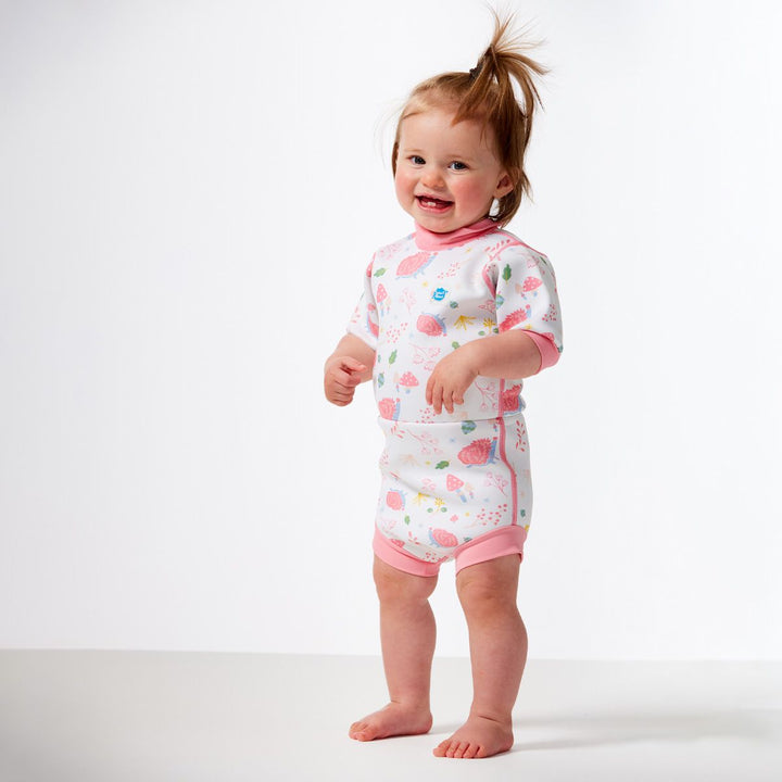 Lifestyle image of child wearing a baby wetsuit with built in swim nappy in white with pink trims and forest themed print, including hedgehogs, mushrooms and leaves. Front.