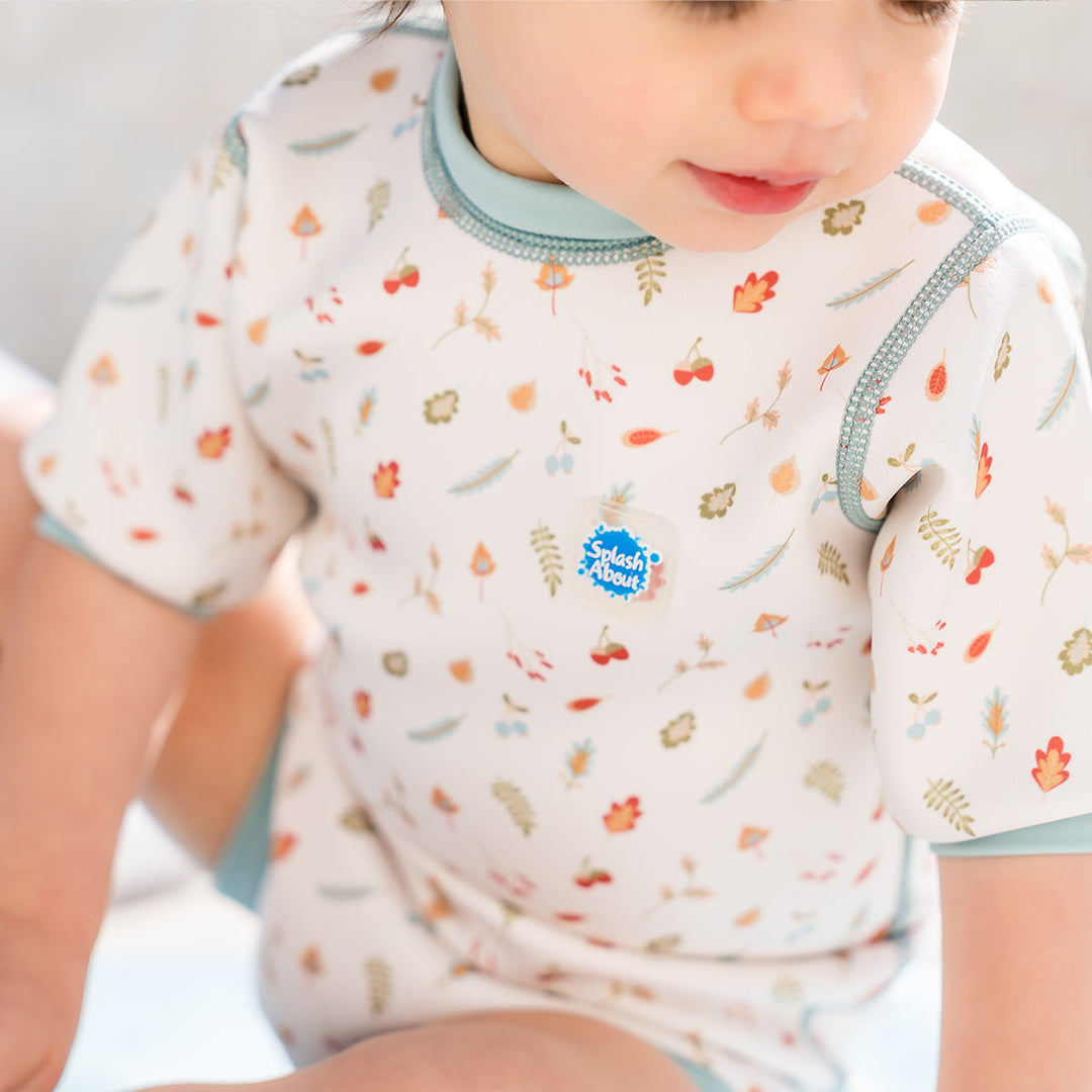 Lifestyle image of child wearing baby wetsuit with built in swim nappy in white with light blue trims and leaves and acorns themed print.