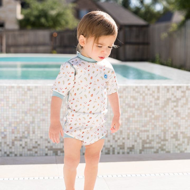 Lifestyle image of child wearing baby wetsuit with built in swim nappy in white with light blue trims and leaves and acorns themed print.