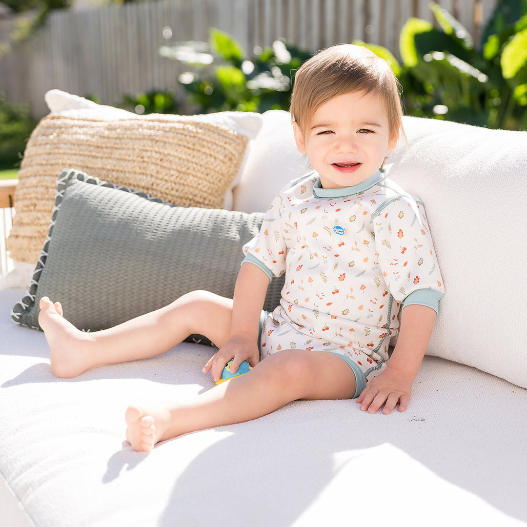 Lifestyle image of child wearing baby wetsuit with built in swim nappy in white with light blue trims and leaves and acorns themed print.