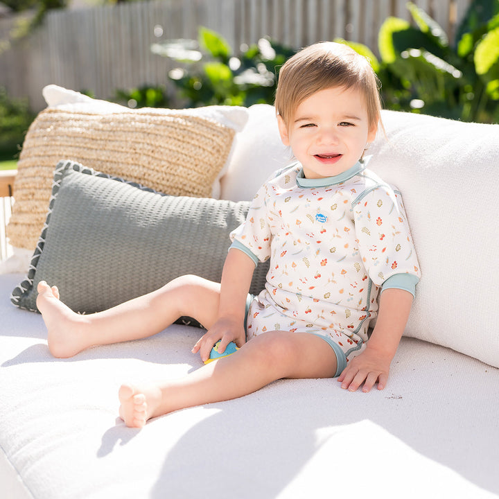 Lifestyle image of child wearing baby wetsuit with built in swim nappy in white with light blue trims and leaves and acorns themed print.