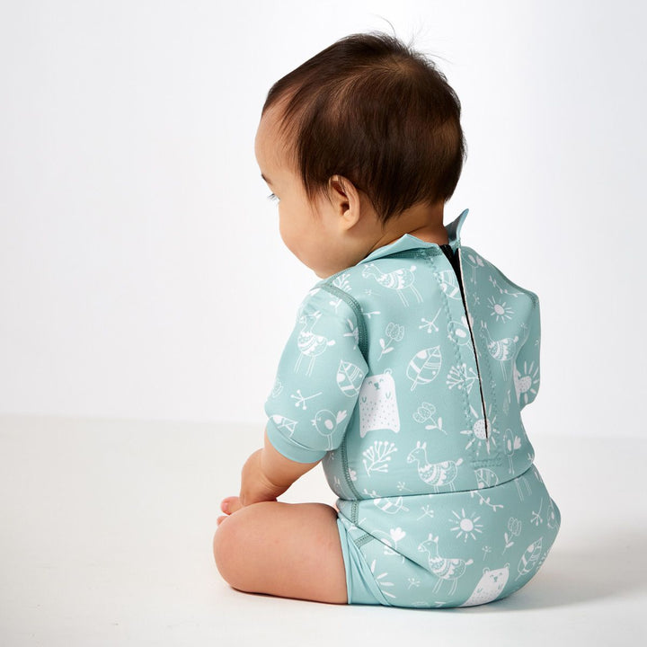 Lifestyle image of child wearing a baby wetsuit with built in swim nappy in greenish blue with animals themed print, including bears, birds and llamas. Back.