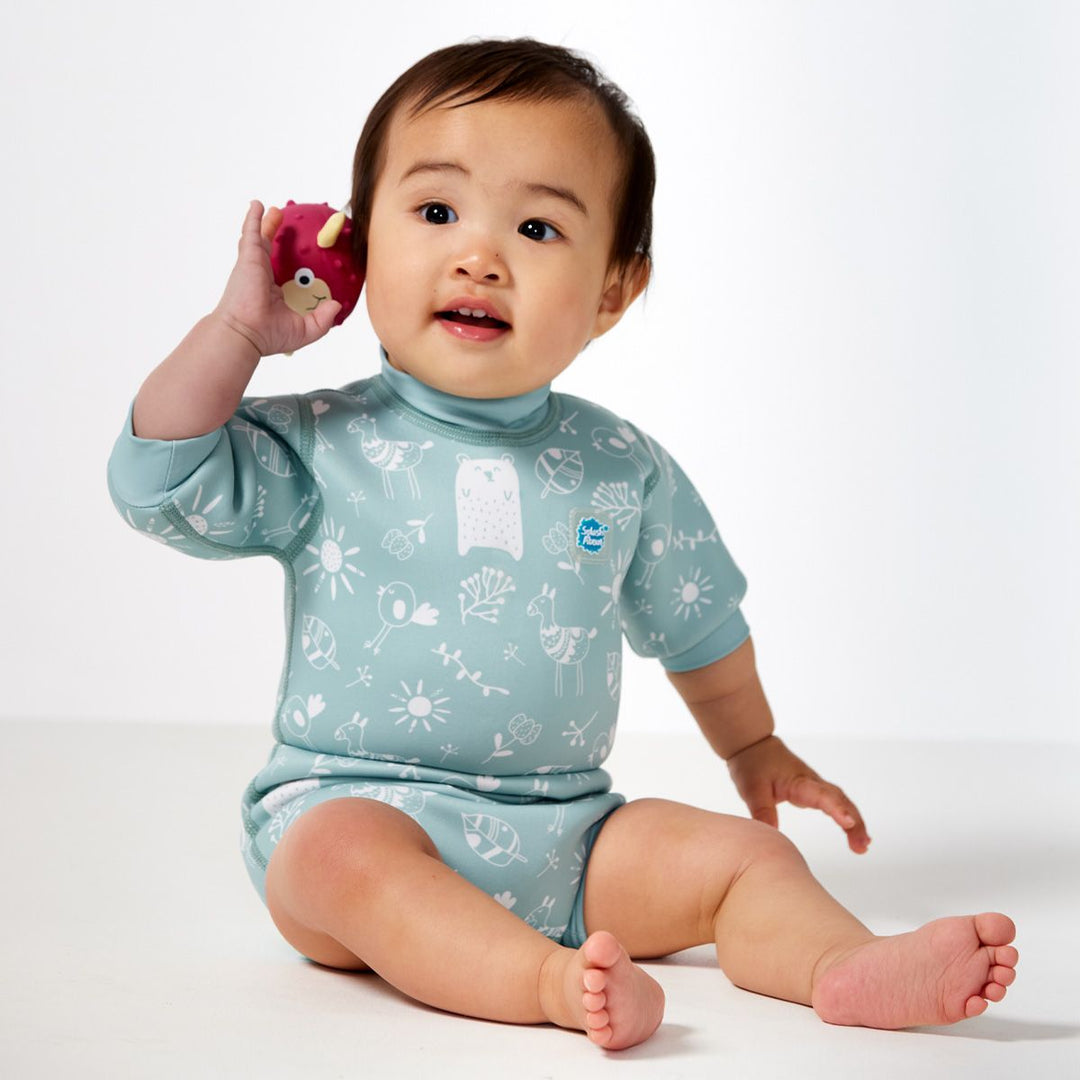 Lifestyle image of child wearing a baby wetsuit with built in swim nappy in greenish blue with animals themed print, including bears, birds and llamas. Front.