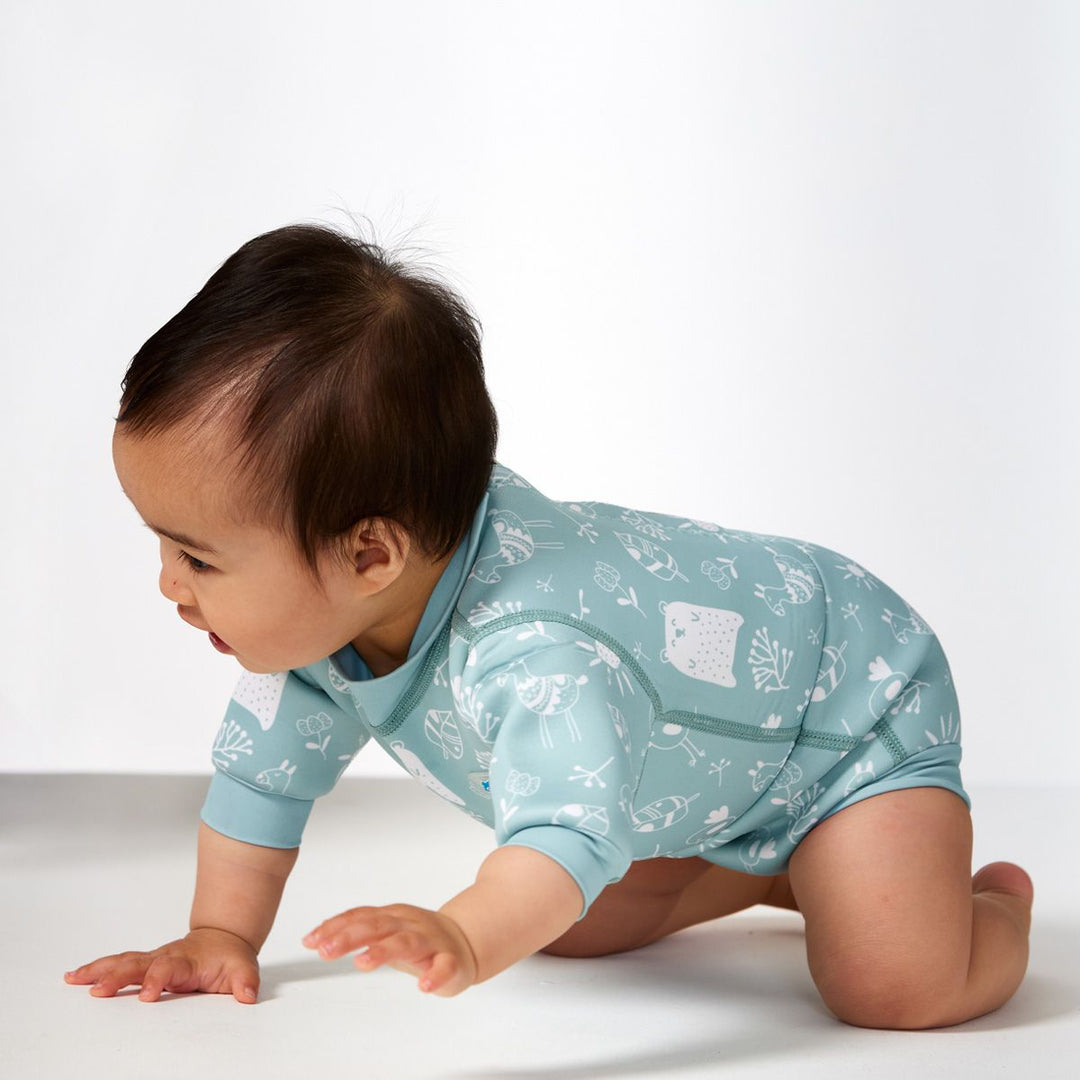 Lifestyle image of child wearing a baby wetsuit with built in swim nappy in greenish blue with animals themed print, including bears, birds and llamas. Side.