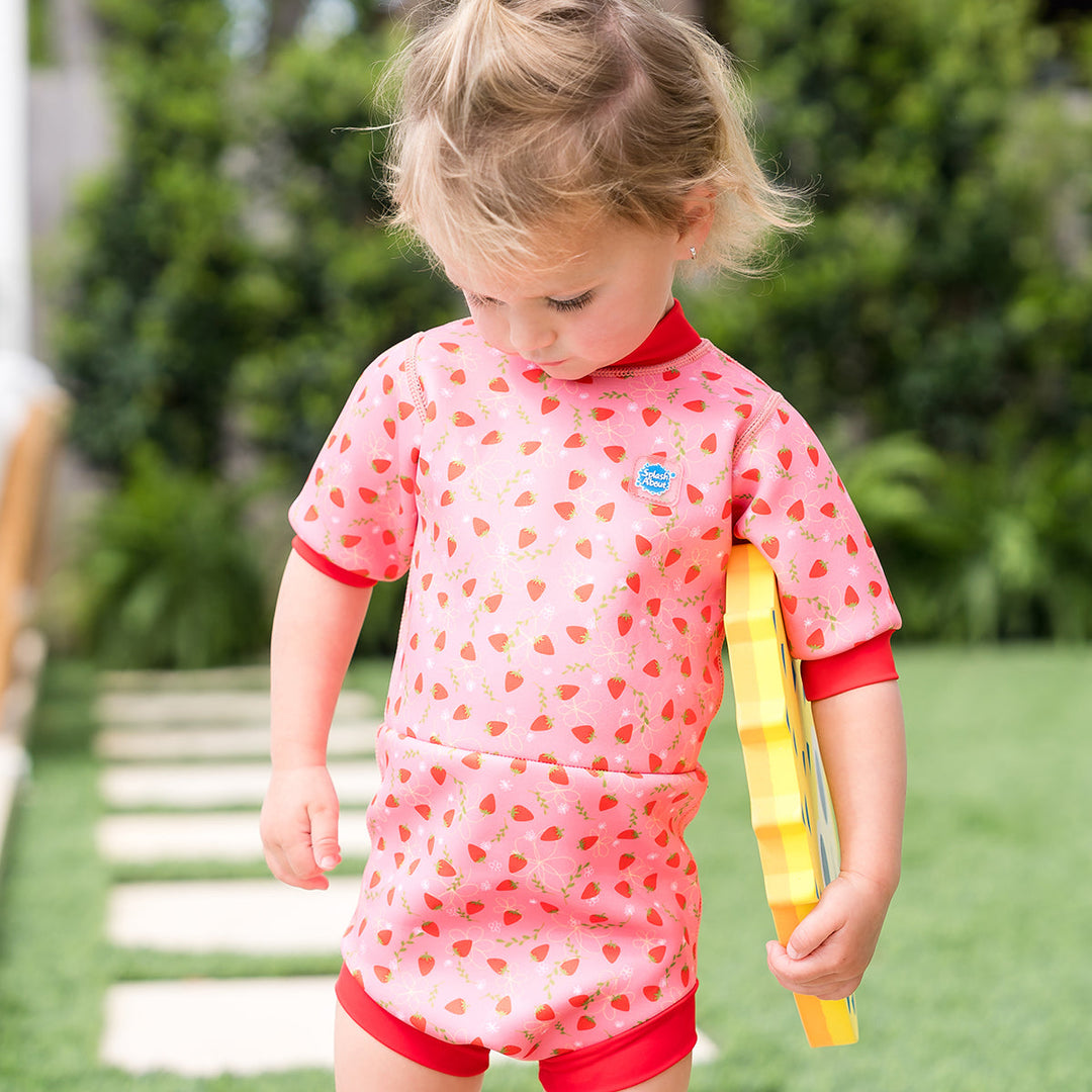 Child wearing baby wetsuit with built in swim nappy in pink with red trims and strawberries themed print.