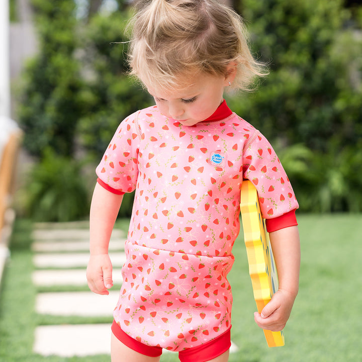 Child wearing baby wetsuit with built in swim nappy in pink with red trims and strawberries themed print.