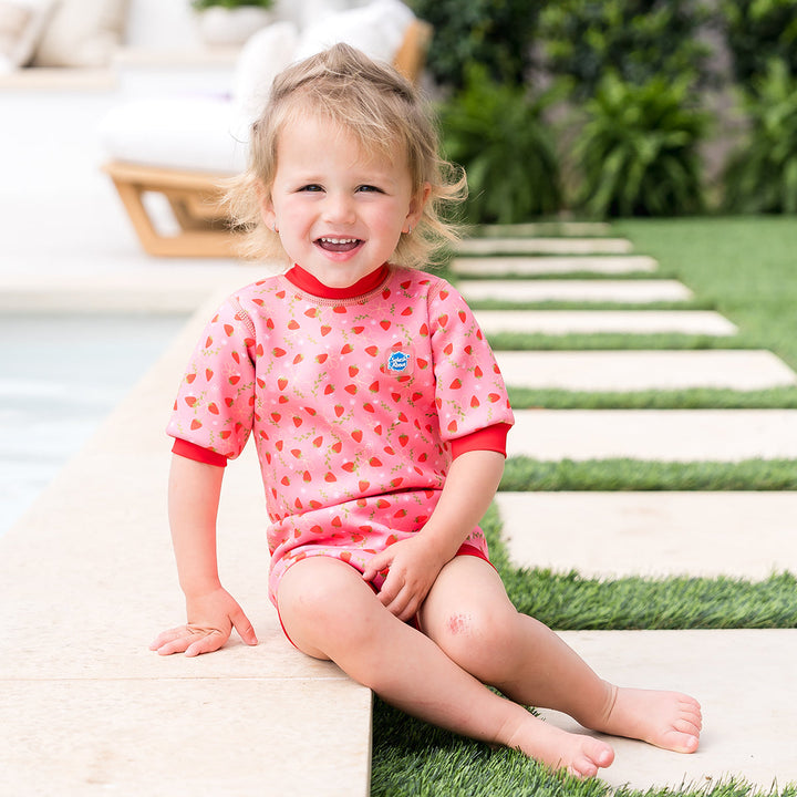Child wearing baby wetsuit with built in swim nappy in pink with red trims and strawberries themed print.