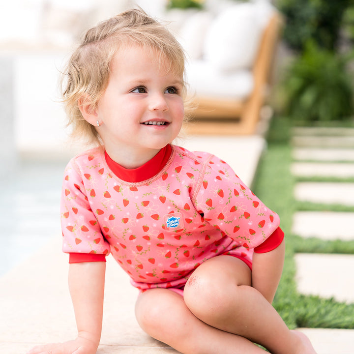 Child wearing baby wetsuit with built in swim nappy in pink with red trims and strawberries themed print.