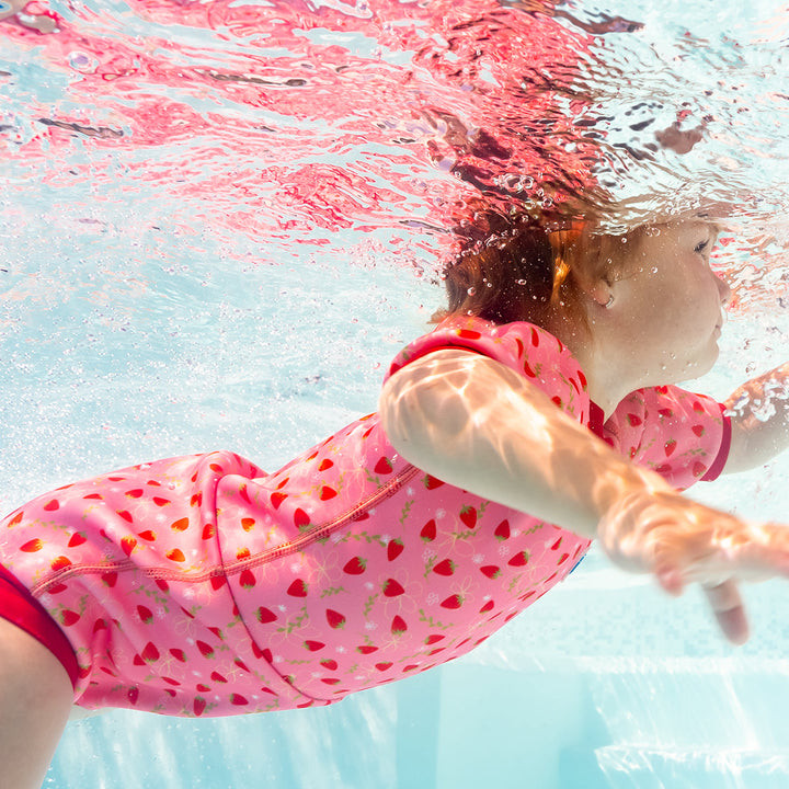 Child wearing baby wetsuit with built in swim nappy in pink with red trims and strawberries themed print.