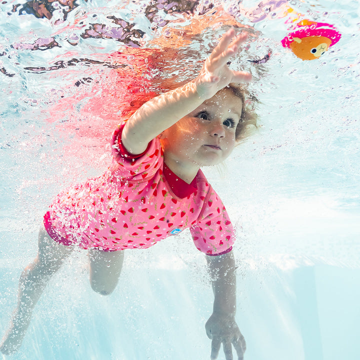 Child wearing baby wetsuit with built in swim nappy in pink with red trims and strawberries themed print.