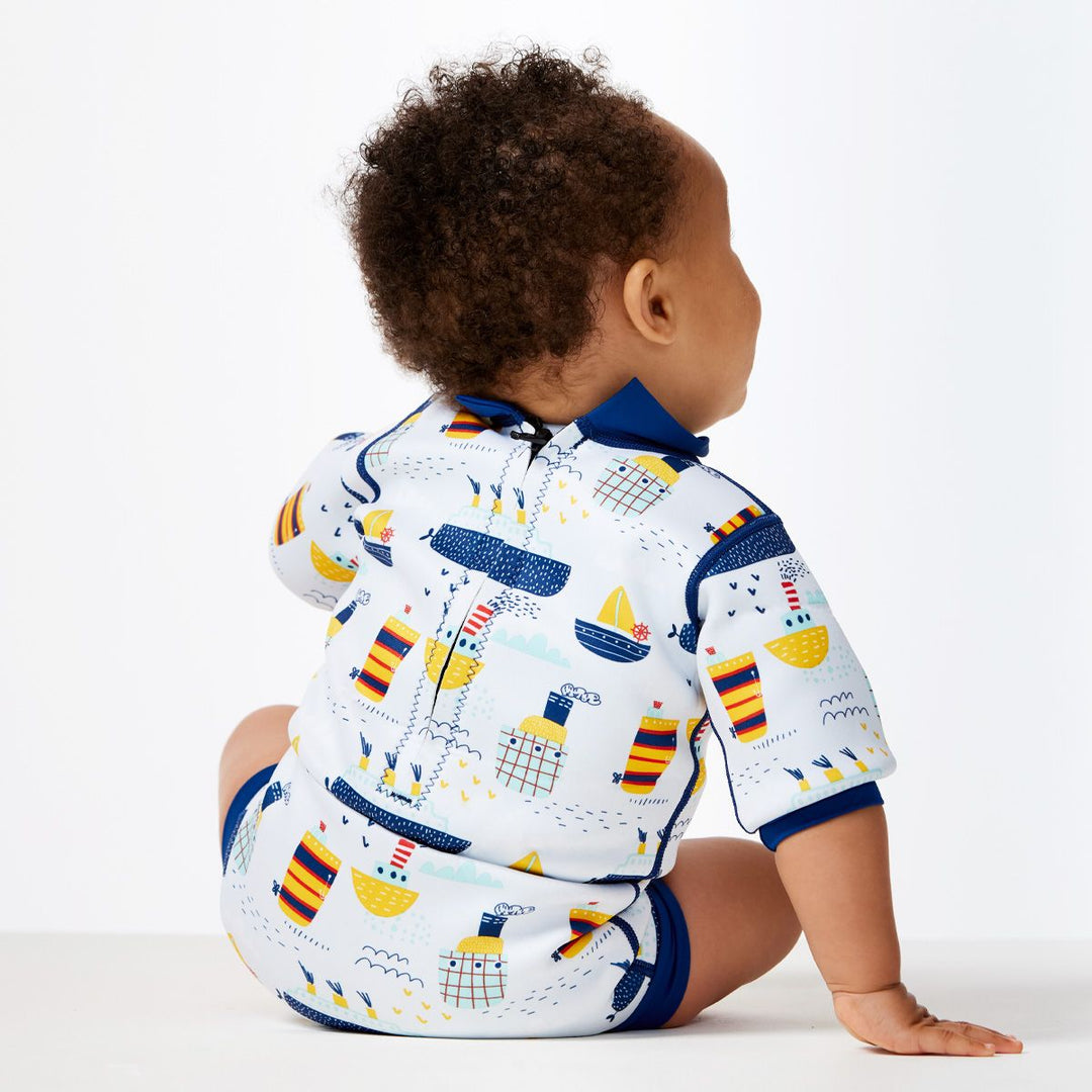 Lifestyle image of child wearing a baby wetsuit with built in swim nappy in white with navy trims and boats themed print. Back.