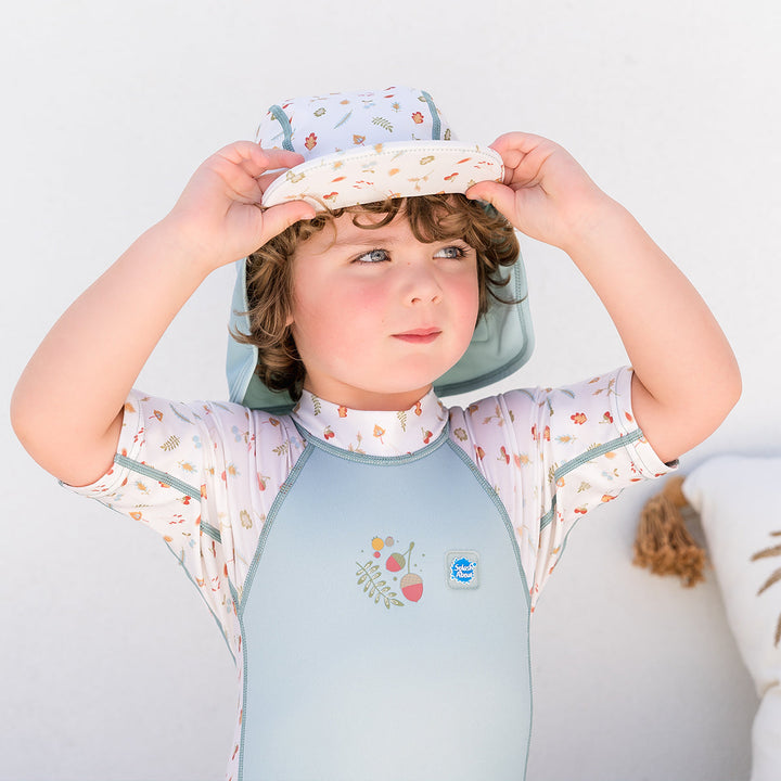 Little one wearing legionnaire style sun hat in light blue and white, with acorns and leaves themed print panel. They are also wearing matching wetsuit.