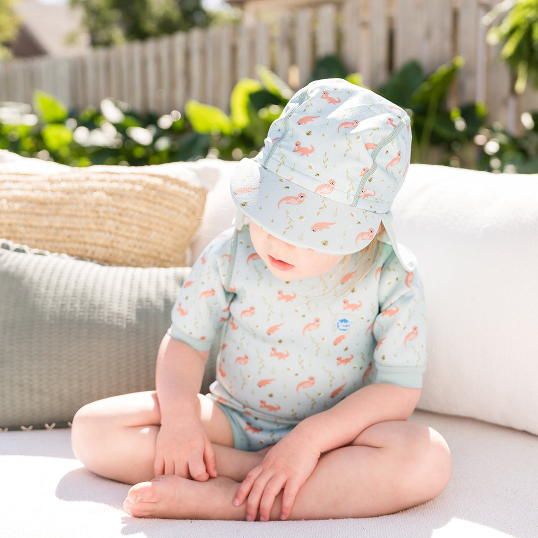 Little one wearing legionnaire style sun hat in light blue, with otters themed print panel. They are also wearing matching wetsuit.
