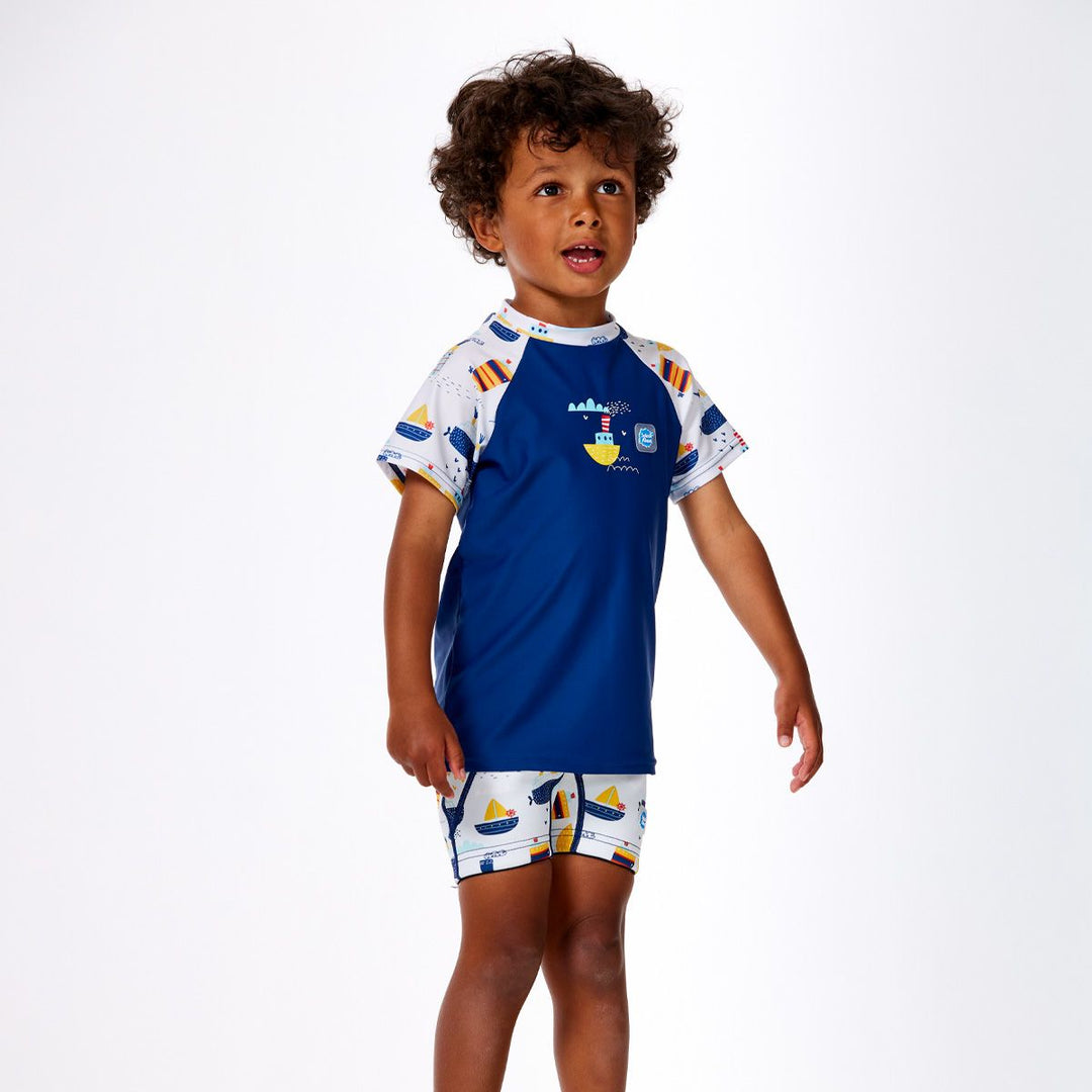 Lifestyle image of child wearing UV protective short sleeve rash top in blue, and boats themed print on the chest and sleeves. He's also wearing matching jammers. Front.