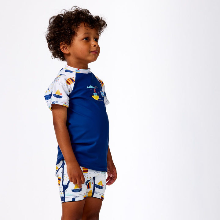 Lifestyle image of child wearing UV protective short sleeve rash top in blue, and boats themed print on the chest and sleeves. He's also wearing matching jammers. Side.
