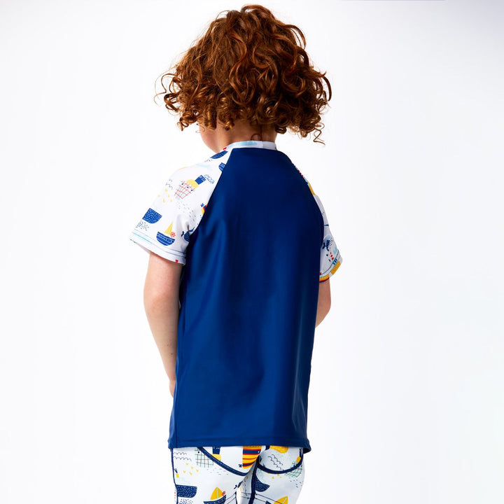 Lifestyle image of child wearing UV protective short sleeve rash top in blue, and boats themed print on the chest and sleeves. He's also wearing matching jammers. Back.
