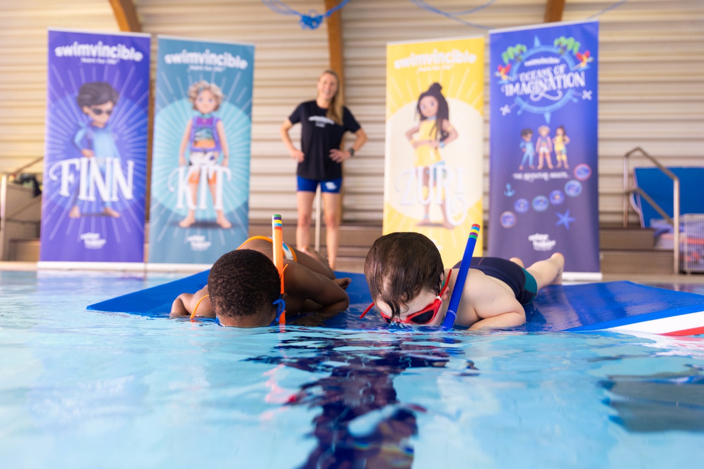 Children in a pool with a swim instructor and promotional banners in the background