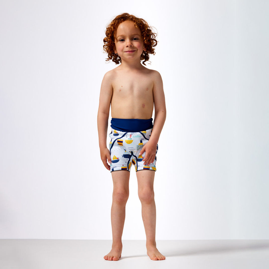 Little one wearing neoprene swim shorts in white with blue navy waist and boats themed print. Front.