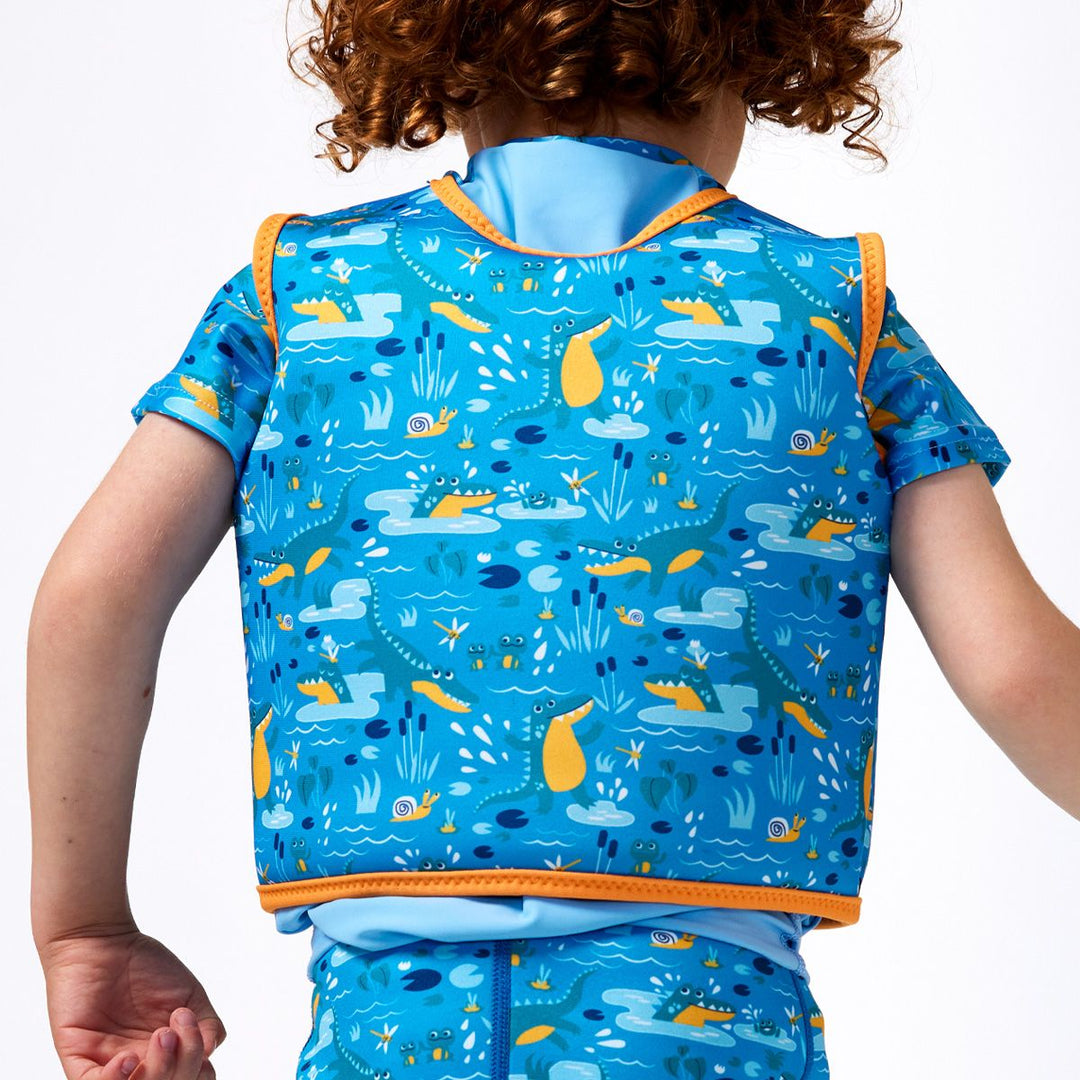 Lifestyle image of child wearing neoprene swim vest for toddlers with non-removable floats in sky blue, orange trims and swamp creatures themed print, including crocodiles and frogs. He's also wearing matching rash top and jammers. Back.