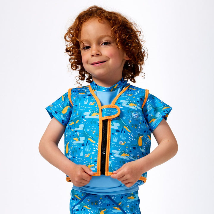 Lifestyle image of child wearing neoprene swim vest for toddlers with non-removable floats in sky blue, orange trims and swamp creatures themed print, including crocodiles and frogs. He's also wearing matching rash top and jammers. Front.