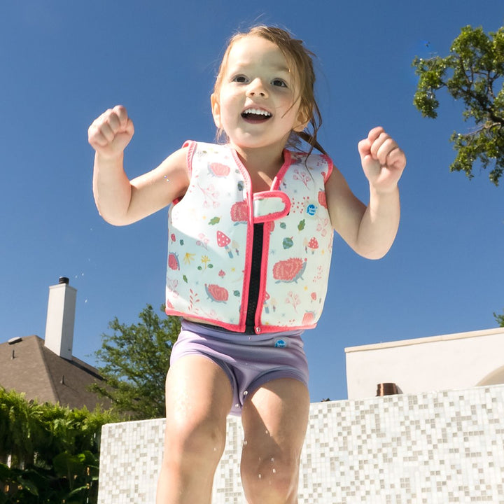 Lifestyle image of child wearing neoprene swim vest for toddlers with non-removable floats in white, pink trims and forest themed print, including hedgehogs, mushrooms and leaves. Front.