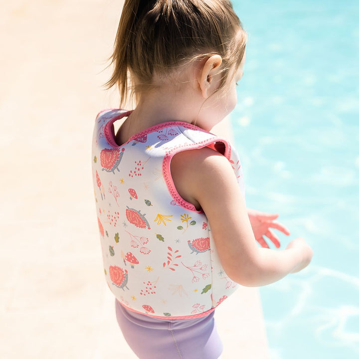 Lifestyle image of child wearing neoprene swim vest for toddlers with non-removable floats in white, pink trims and forest themed print, including hedgehogs, mushrooms and leaves. Back.
