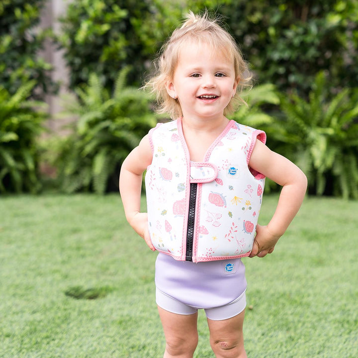 Lifestyle image of child wearing neoprene swim vest for toddlers with non-removable floats in white, pink trims and forest themed print, including hedgehogs, mushrooms and leaves. Front.