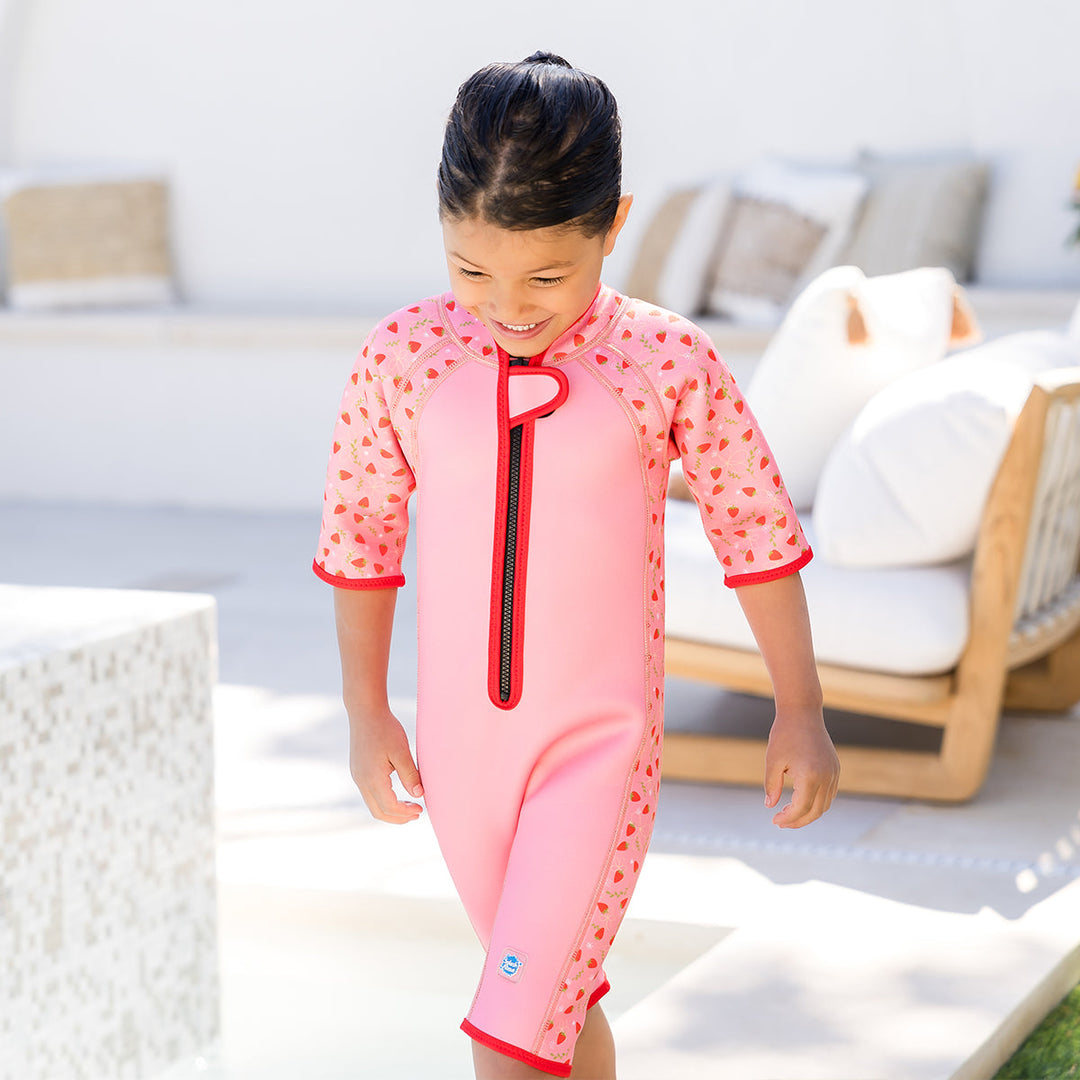 Girl wearing shorty wetsuit in pink with bright red trims, featuring strawberries themed print in sleeves and side panels. She's also wearing matching legionnaire hat.