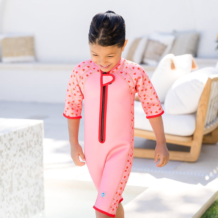 Girl wearing shorty wetsuit in pink with bright red trims, featuring strawberries themed print in sleeves and side panels. She's also wearing matching legionnaire hat.