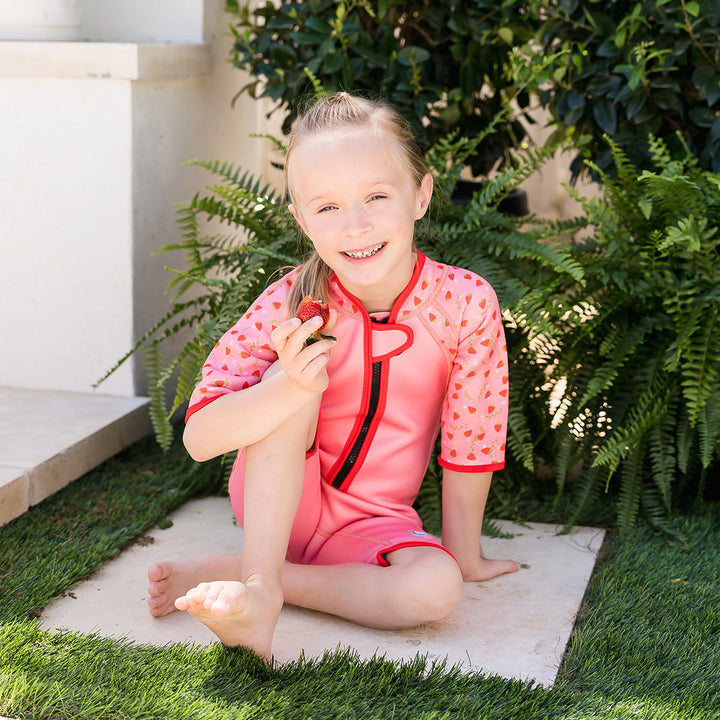 Girl wearing shorty wetsuit in pink with bright red trims, featuring strawberries themed print in sleeves and side panels.