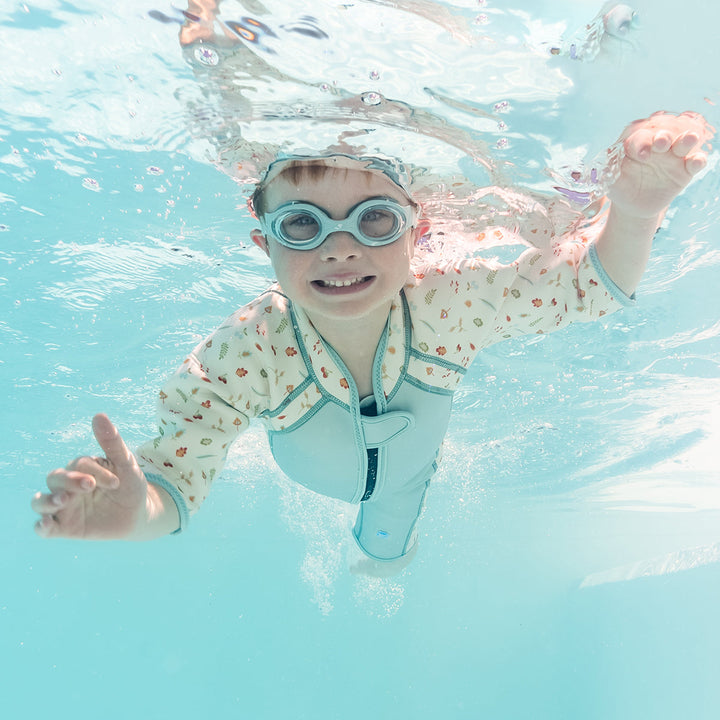 Kid swimming in shorty wetsuit in light blue, featuring acorns and leaves themed print over white background in sleeves and side panels. He's also wearing blue goggles.