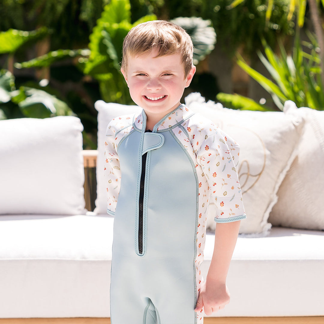 Kid wearing shorty wetsuit in light blue, featuring acorns and leaves themed print over white background in sleeves and side panels. 