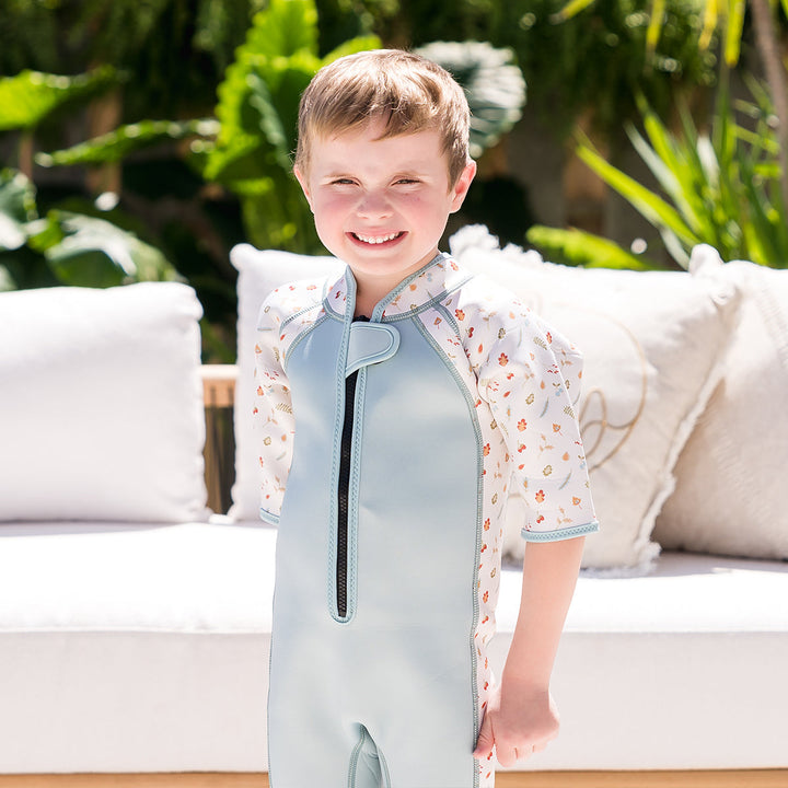 Kid wearing shorty wetsuit in light blue, featuring acorns and leaves themed print over white background in sleeves and side panels. 