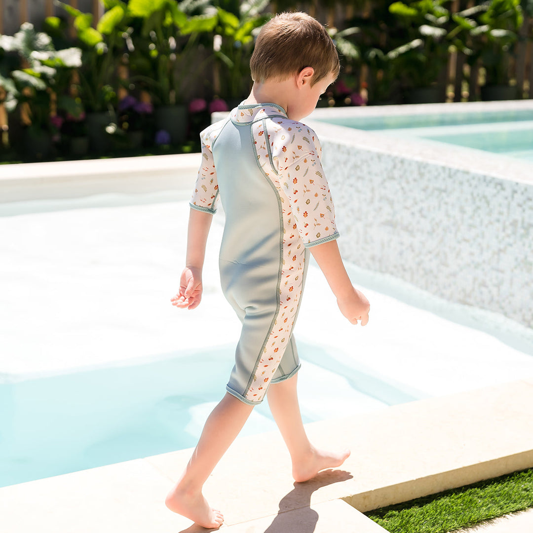 Kid wearing shorty wetsuit in light blue, featuring acorns and leaves themed print over white background in sleeves and side panels. 