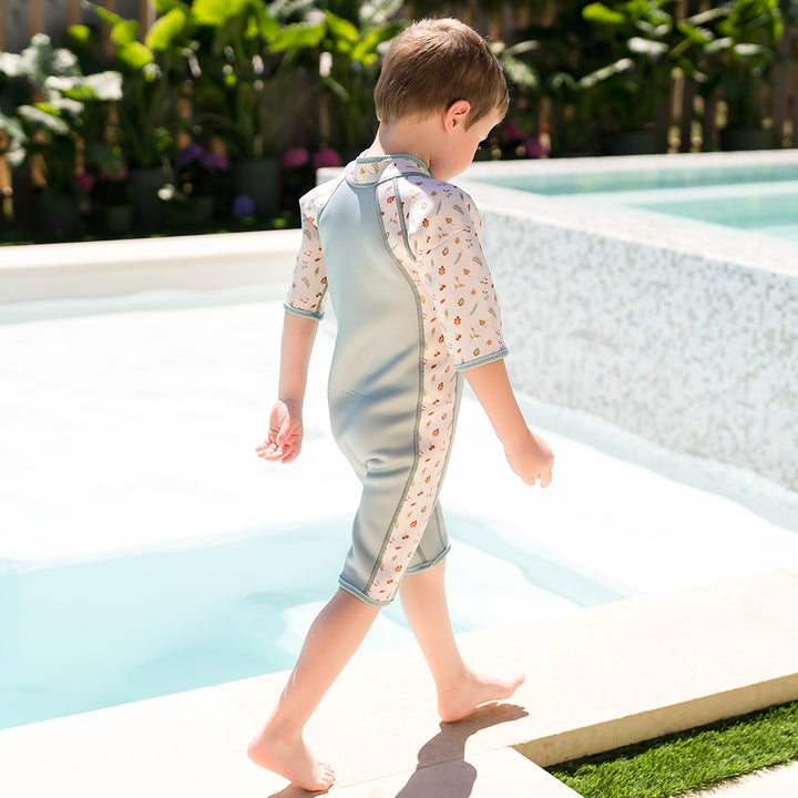Kid wearing shorty wetsuit in light blue, featuring acorns and leaves themed print over white background in sleeves and side panels. 