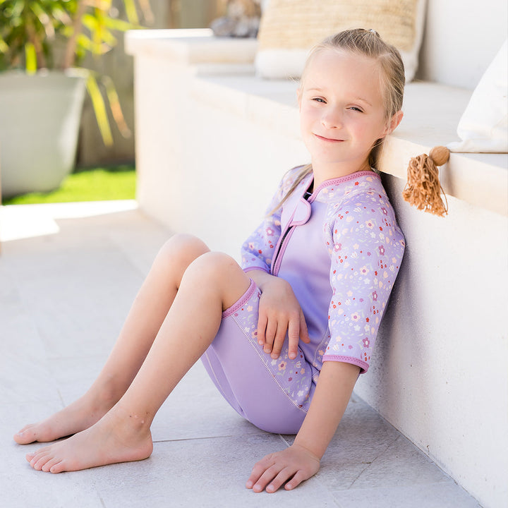 Kid wearing shorty wetsuit in lilac, featuring floral themed print in sleeves and side panels.