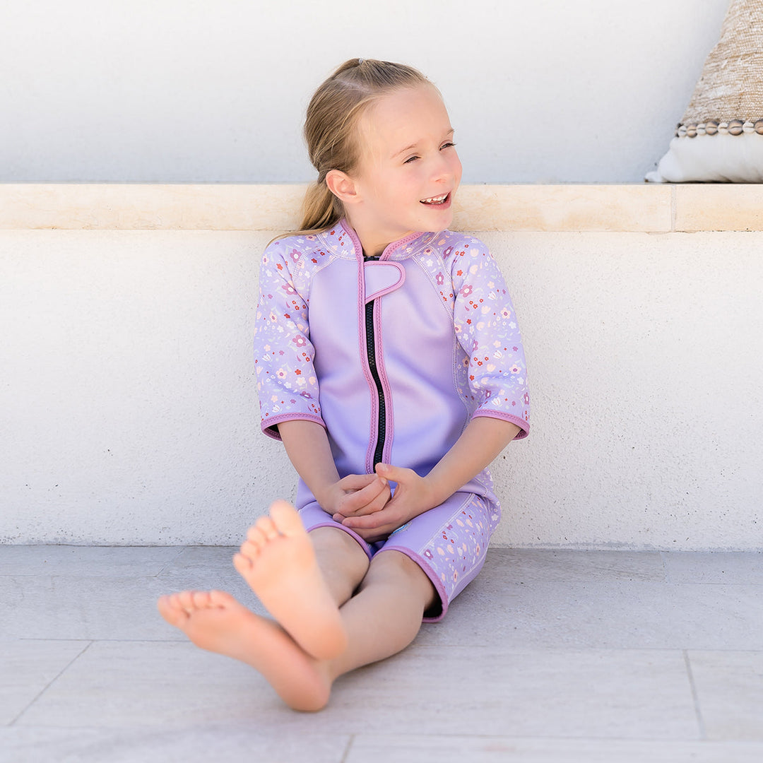 Kid wearing shorty wetsuit in lilac, featuring floral themed print in sleeves and side panels.