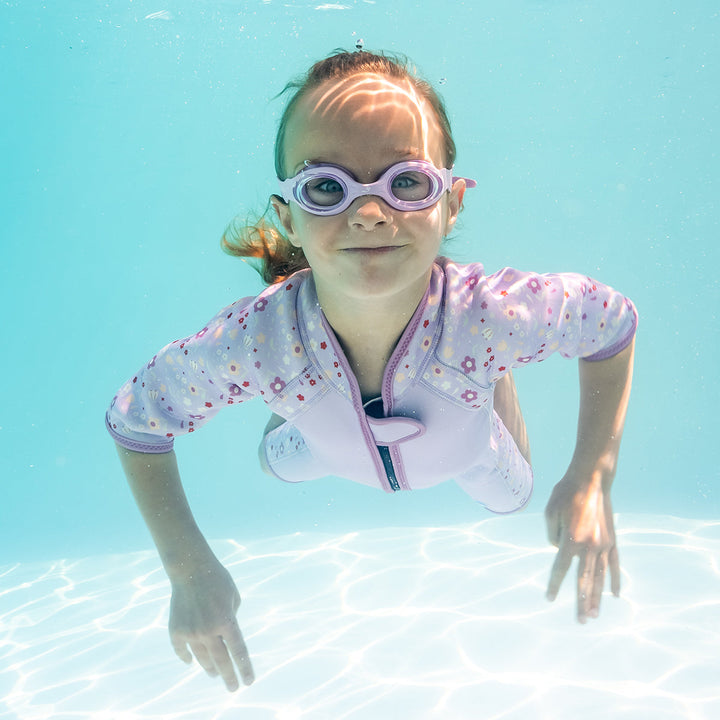Kid swimming in shorty wetsuit in lilac, featuring floral themed print in sleeves and side panels. She is also wearing lilac goggles.