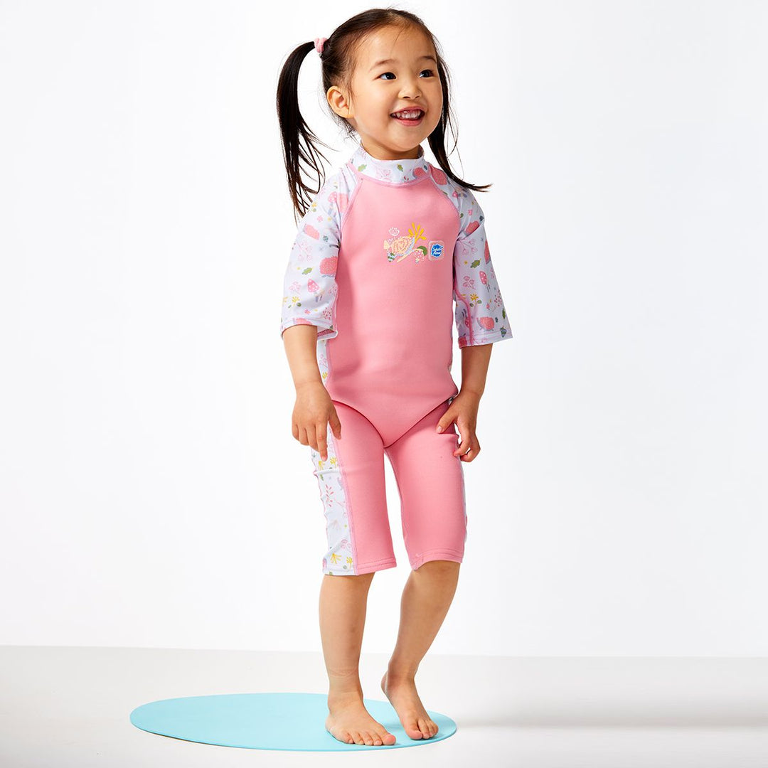 Lifestyle image of child wearing one piece UV sun and sea wetsuit for toddlers in pink. Forest themed print including hedgehogs, mushrooms and leaves on sleeves, side panels, neck and chest. Front.