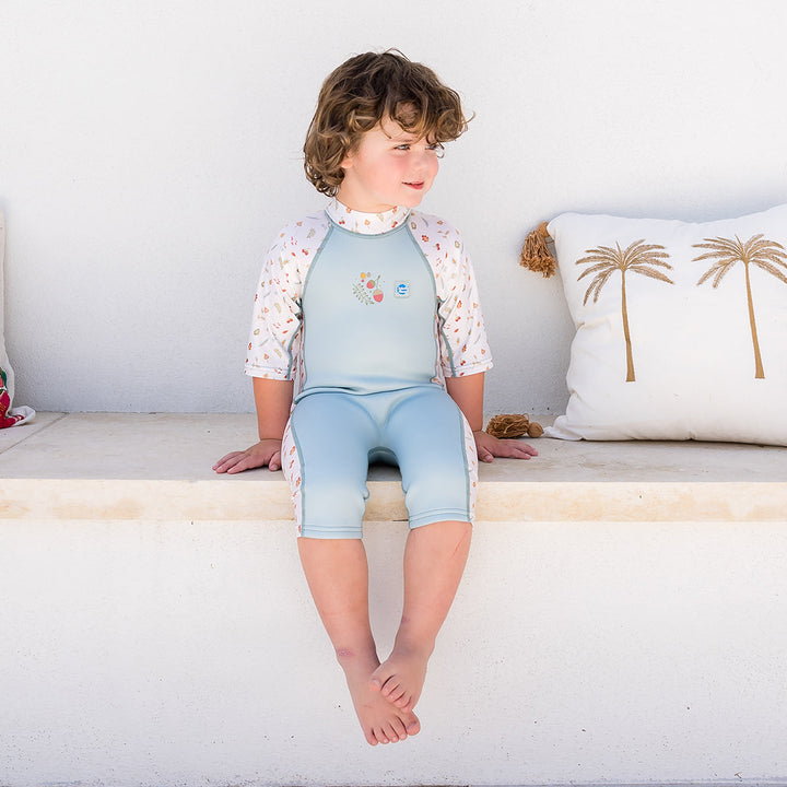 Child wearing one piece UV sun and sea wetsuit for toddlers in white and light blue. Acorns and leaves themed print on sleeves, side panels, neck and chest.
