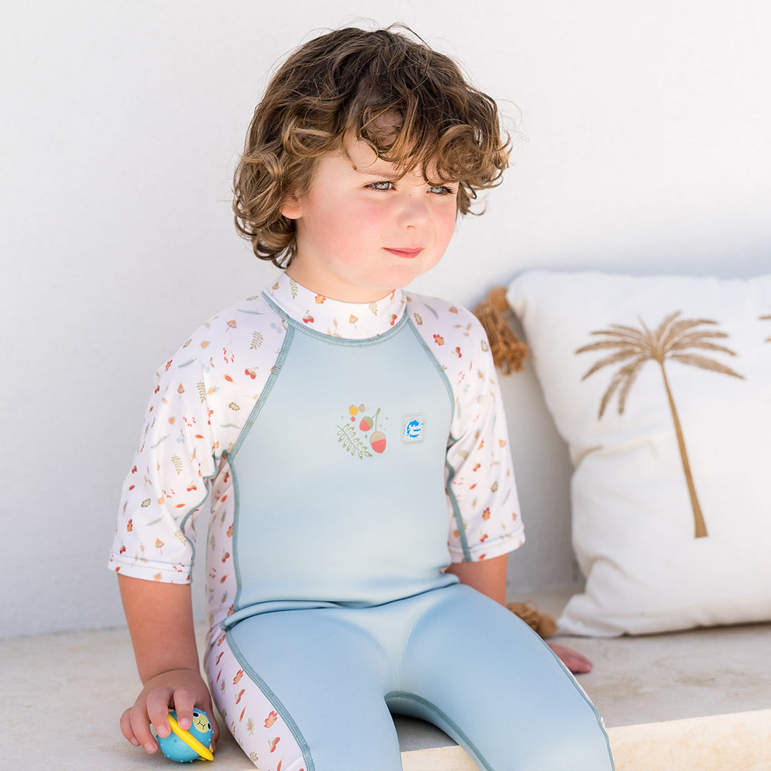 Child wearing one piece UV sun and sea wetsuit for toddlers in white and light blue. Acorns and leaves themed print on sleeves, side panels, neck and chest.