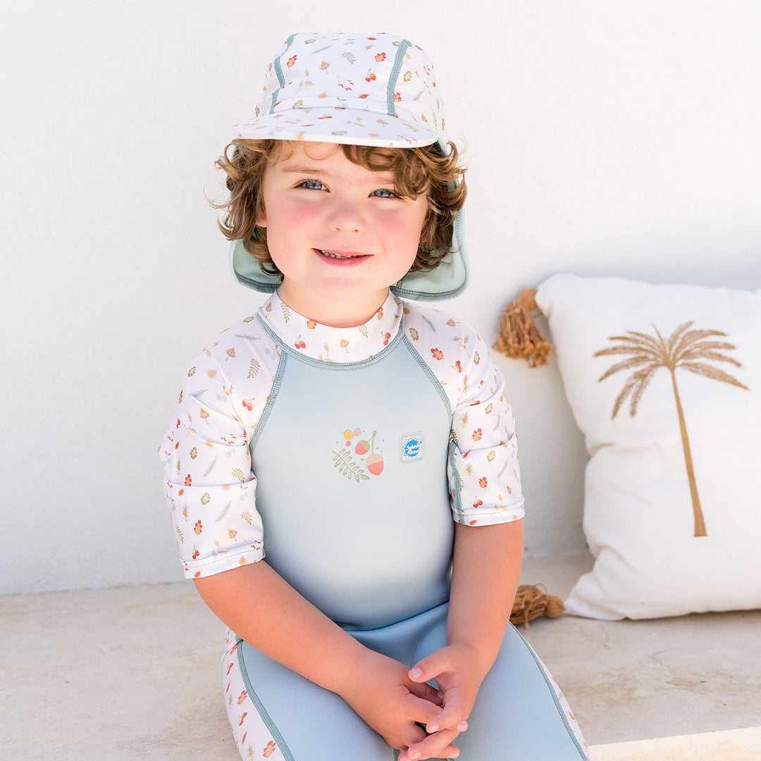 Child wearing one piece UV sun and sea wetsuit for toddlers in white and light blue. Acorns and leaves themed print on sleeves, side panels, neck and chest. Child is also wearing matching legionnaire hat.