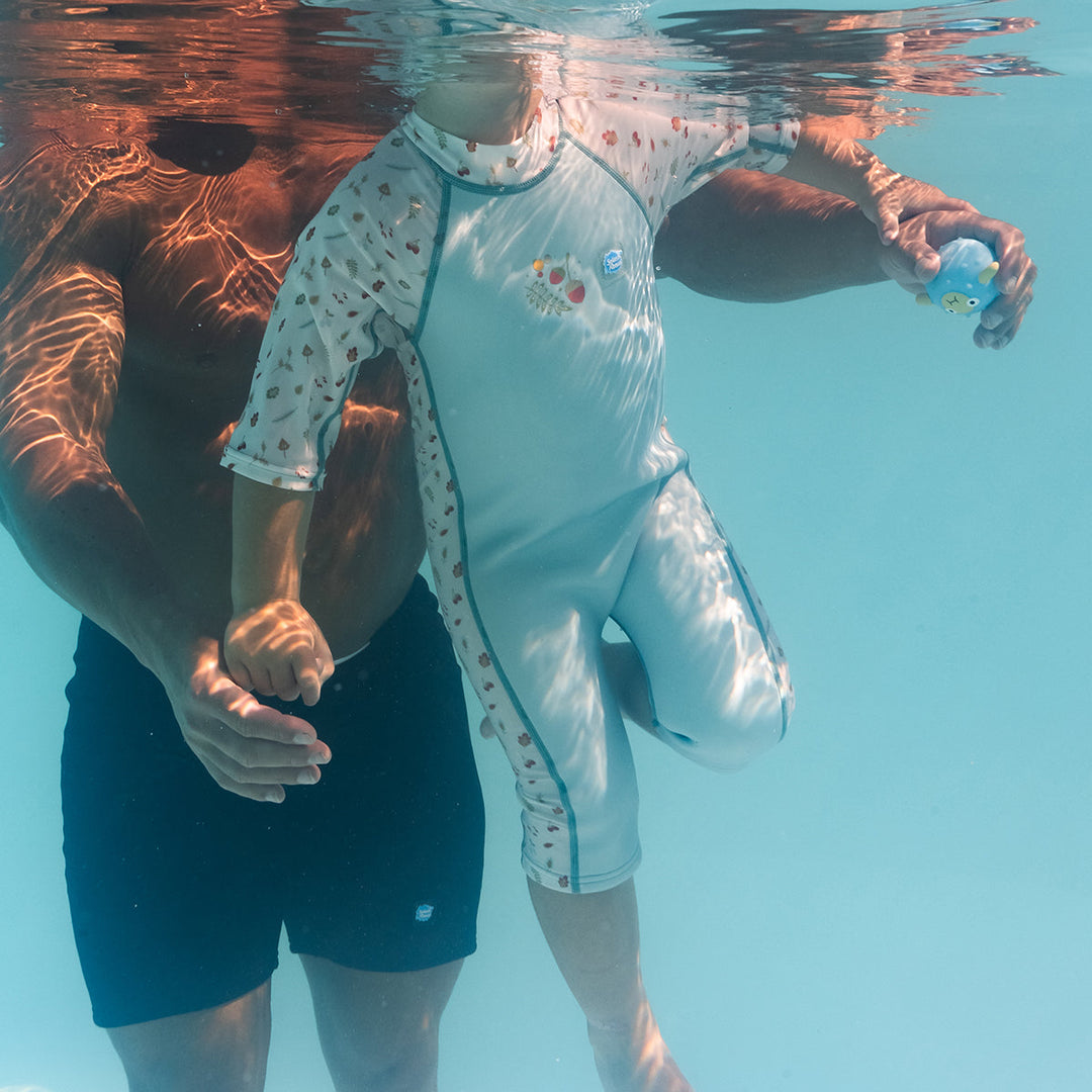 Child swimming in a one piece UV sun and sea wetsuit for toddlers in white and light blue, poolside. Acorns and leaves themed print on sleeves, side panels, neck and chest.