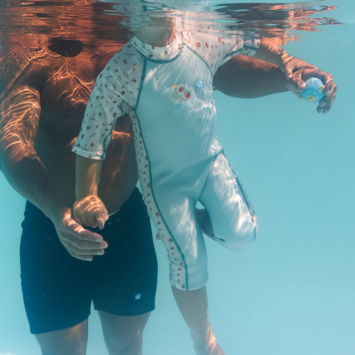 Child swimming in a one piece UV sun and sea wetsuit for toddlers in white and light blue, poolside. Acorns and leaves themed print on sleeves, side panels, neck and chest.