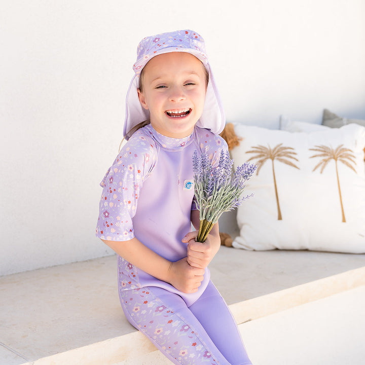 Child wearing oe piece UV sun and sea wetsuit for toddlers in lilac featuring floral print on sleeves, side panels, neck and chest. Child is also wearing matching legionnaire hat.