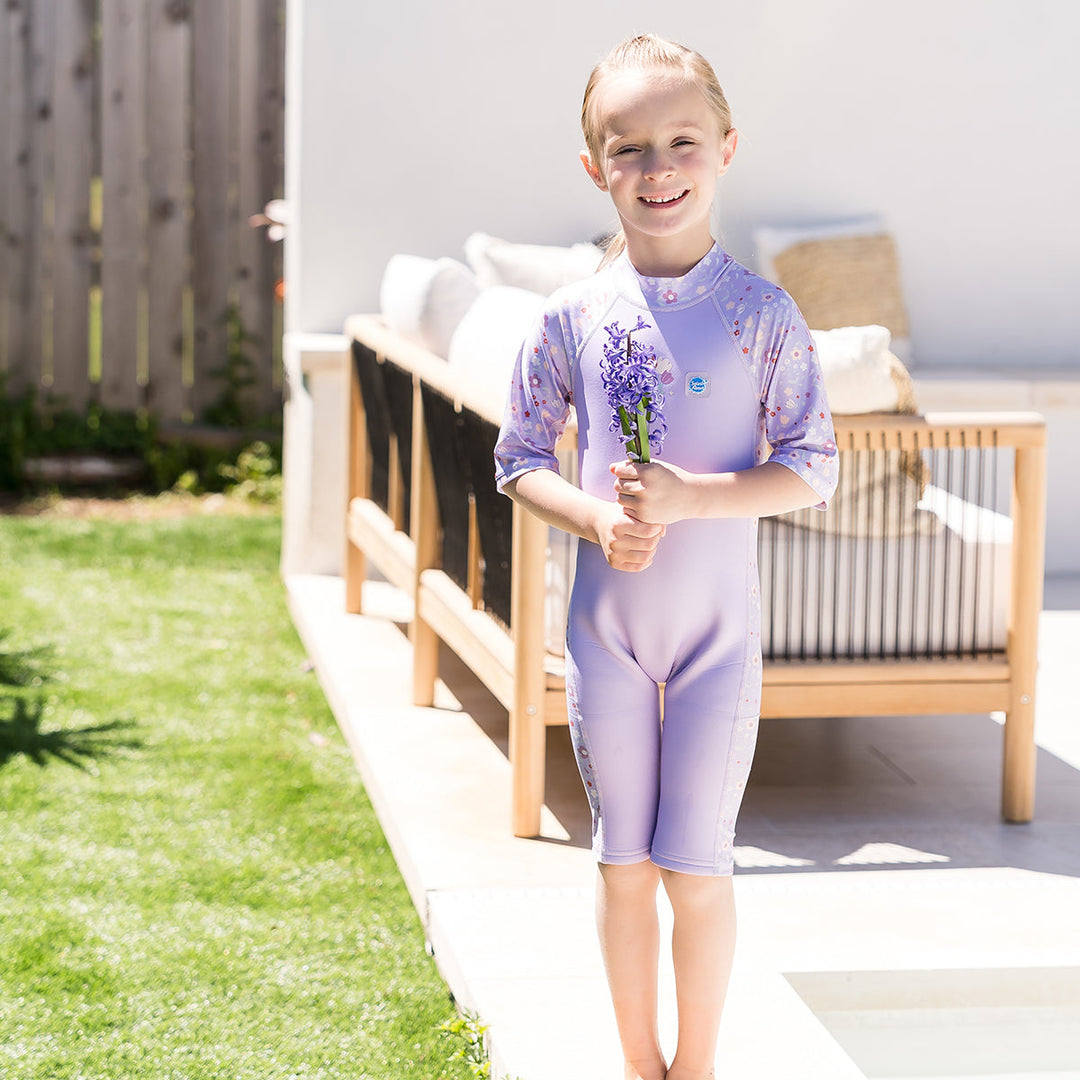 Child wearing oe piece UV sun and sea wetsuit for toddlers in lilac featuring floral print on sleeves, side panels, neck and chest.