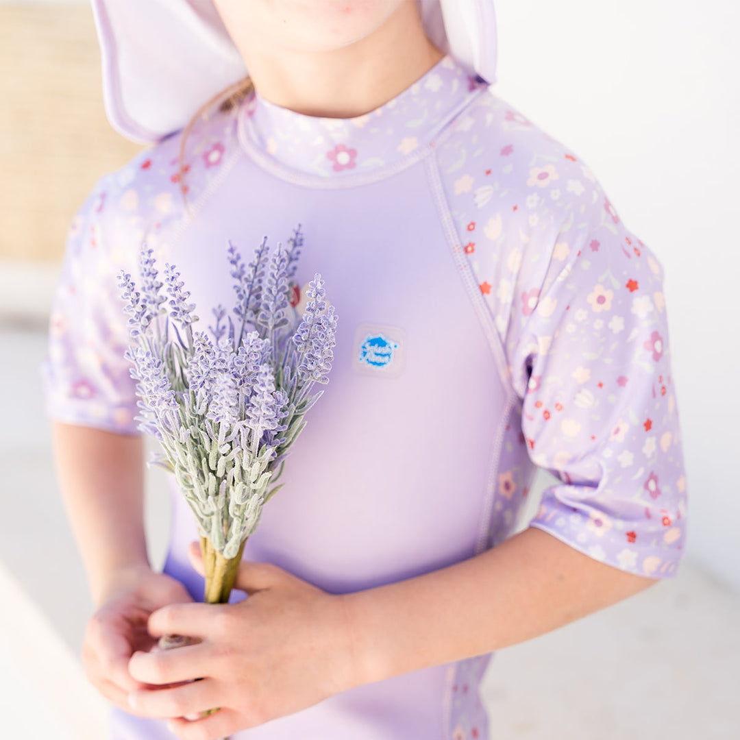 Close-up of child wearing oe piece UV sun and sea wetsuit for toddlers in lilac featuring floral print on sleeves, side panels, neck and chest, holding lilacs.
